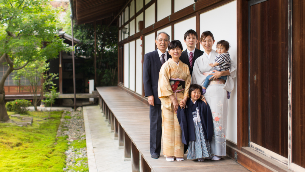 A Japanese family posing on the wooden engawa (veranda) of a traditional house. The group consists of three generations, with older members in formal attire and younger members wearing traditional kimono. The background features a beautiful Japanese garden with lush greenery.