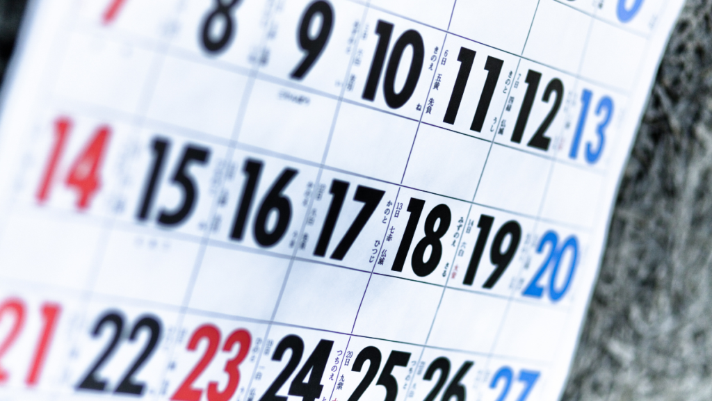 A close-up of a Japanese calendar showing dates in black and red, with Japanese characters indicating holidays and special days. The calendar is slightly tilted, with a blurred background, highlighting the focus on the numbers and text.