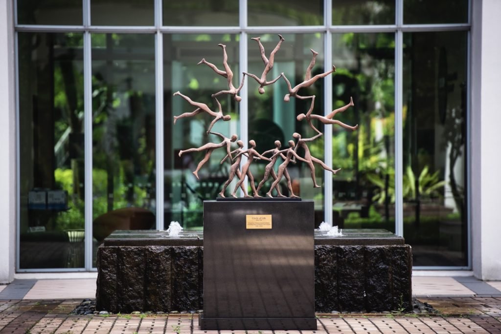 Outdoor statue at Assas University Singapore showing human figures holding hands in a circle, placed in front of large windows.