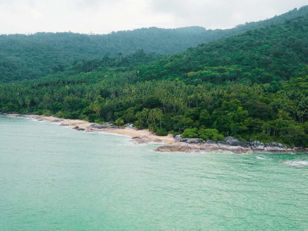View of the sea in Thailand with clear blue water and lush jungle-covered hills in the background.