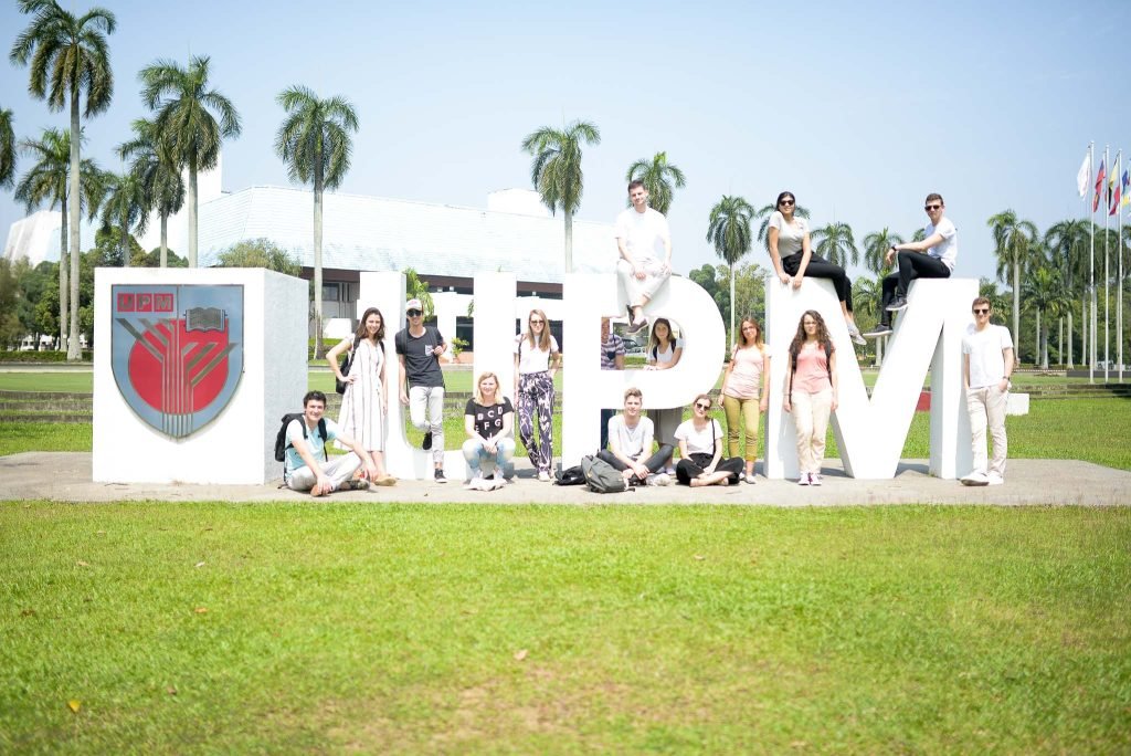 A group of people poses around large white letters "UPM" on a sunny day, with palm trees and flags in the background. The mood is cheerful and relaxed.