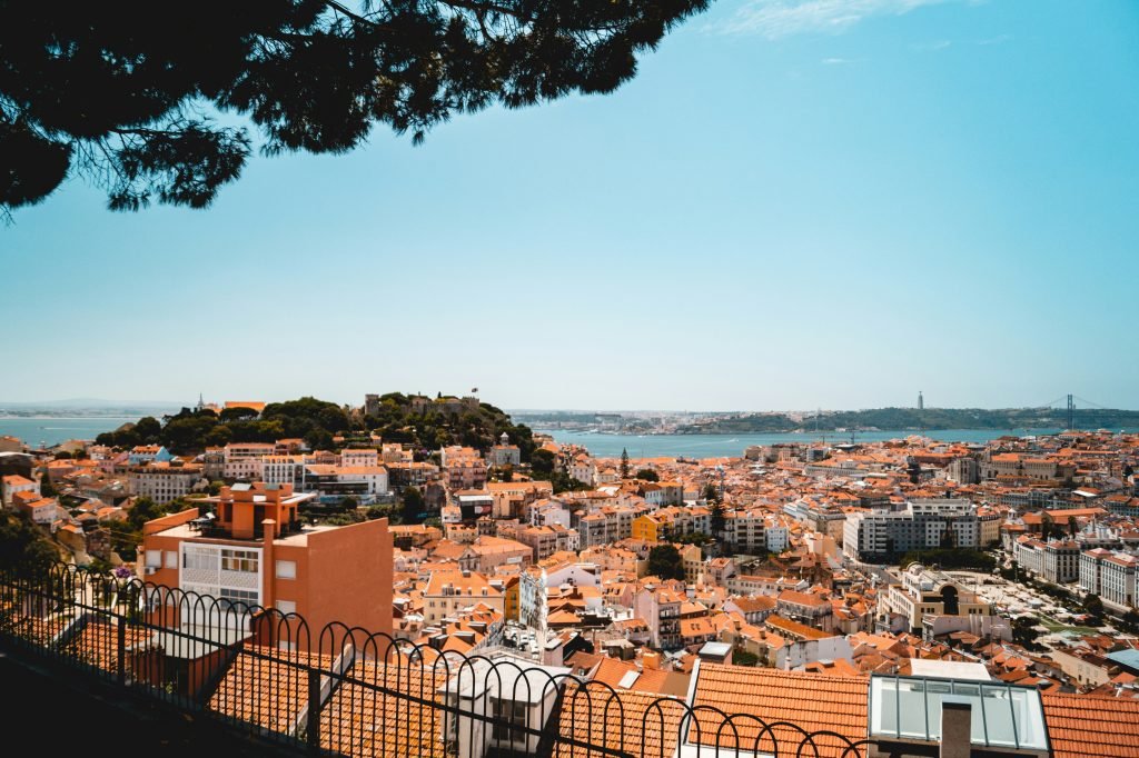 Hillside view of Lisbon, Portugal, with terracotta rooftops, colorful buildings, and a bright blue sky stretching over the historic cityscape.
