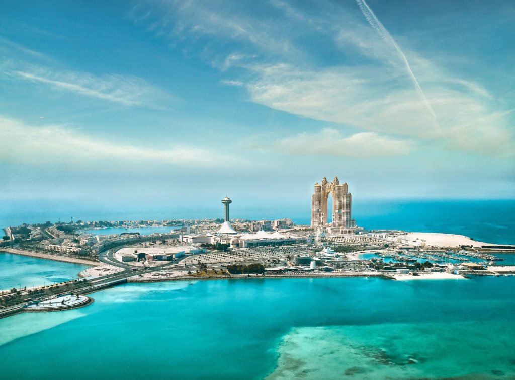 Aerial view of Dubai’s coastal area with turquoise waters in the foreground and the luxurious Emirates Palace and Marina Mall complex on the peninsula. The scene features modern towers, wide roads, and a bright blue sky with streaks of clouds and a visible contrail.