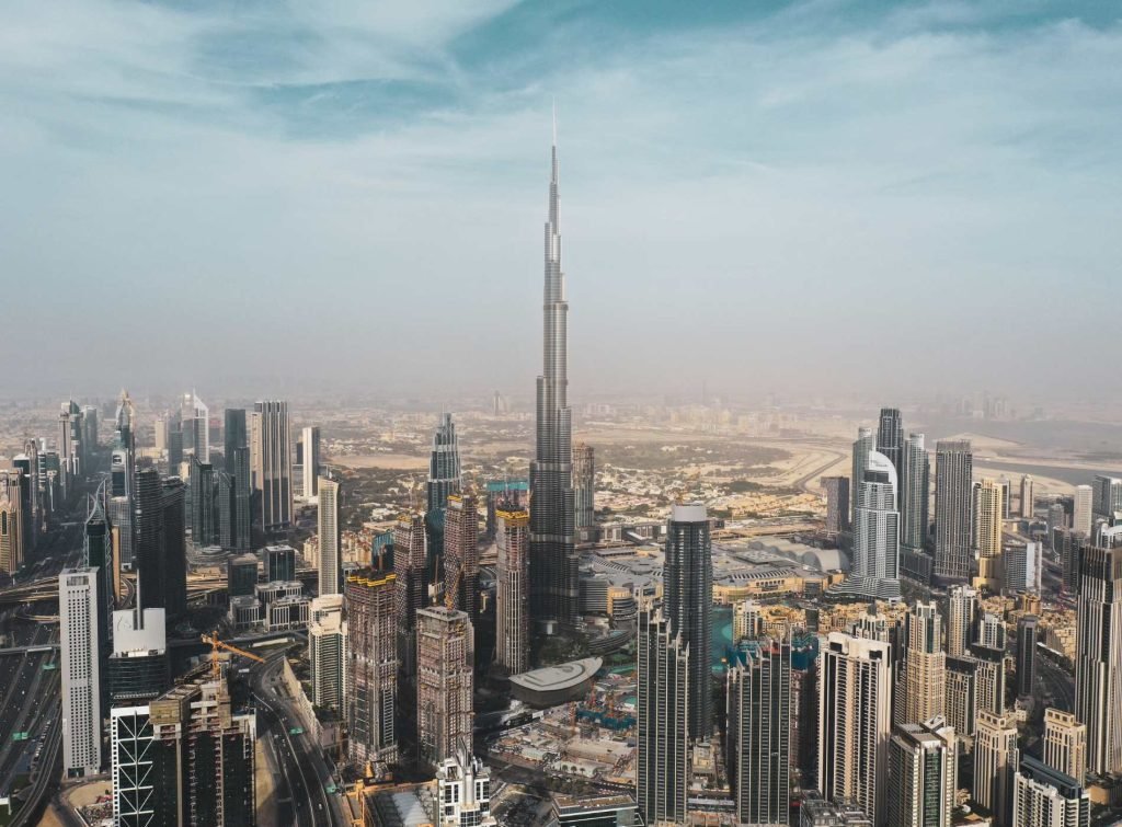 Daytime aerial view of downtown Dubai featuring the Burj Khalifa rising prominently above the dense cityscape of high-rise buildings and construction sites, under a partly cloudy sky. The image highlights the urban density and vertical architecture of the city.
