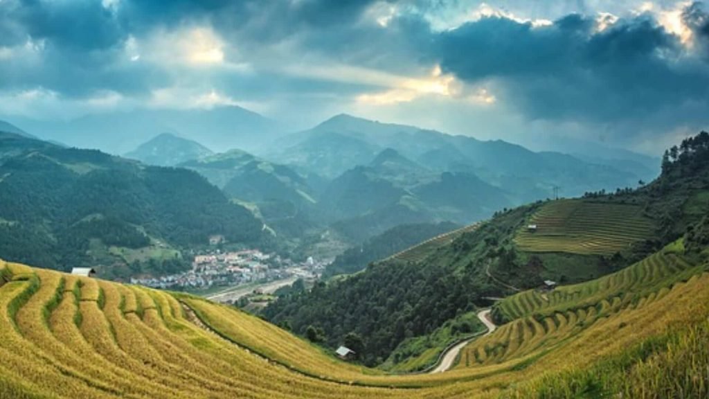 Mountainous landscape with golden terraced fields leading to a village in a valley. Dramatic clouds and sunlight create a serene, majestic atmosphere.