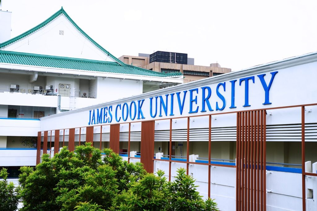 Exterior view of James Cook University Singapore with bold blue signage, white facade, and greenery in the foreground under a bright sky.