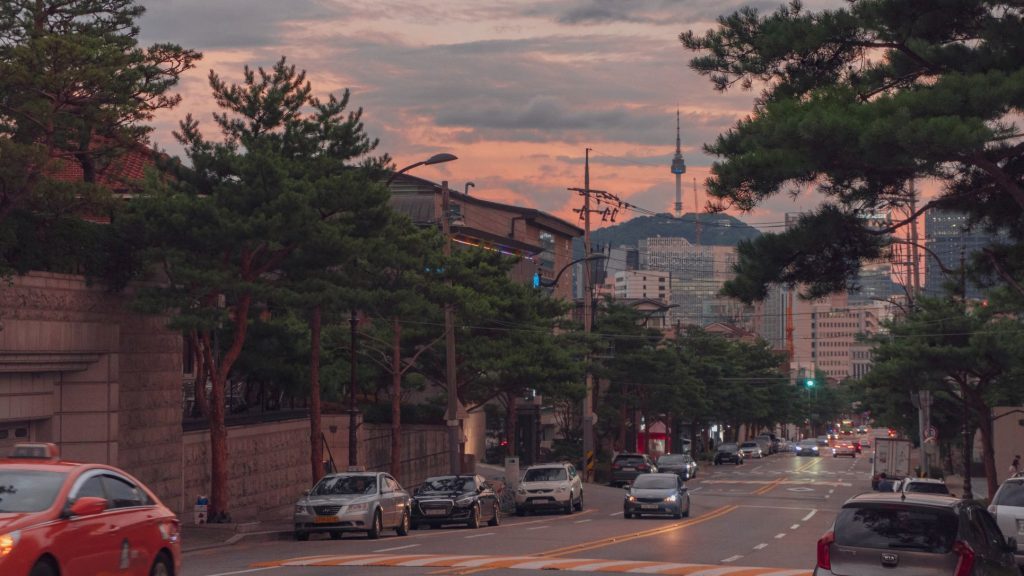 Street lined with cars and trees at dusk, with a cityscape and tower in the background. The sky is a warm, soft pink, evoking a calm, serene mood.