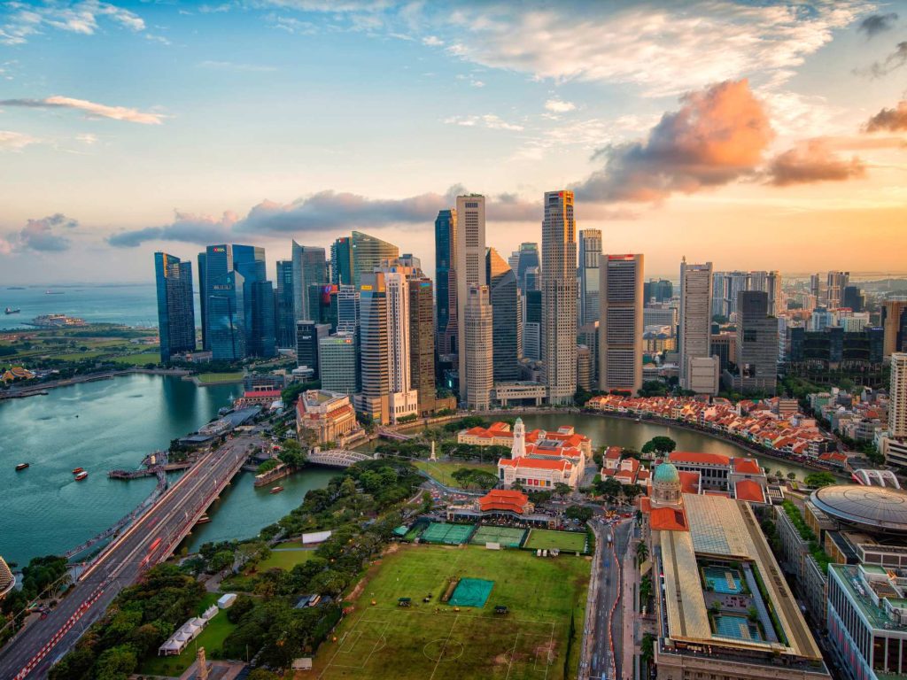 Aerial view of Singapore’s central business district at sunset, showcasing towering skyscrapers, the Singapore River, and iconic red-roofed colonial buildings below. The scene captures the contrast between lush green fields in the foreground and the densely built urban skyline in the background.