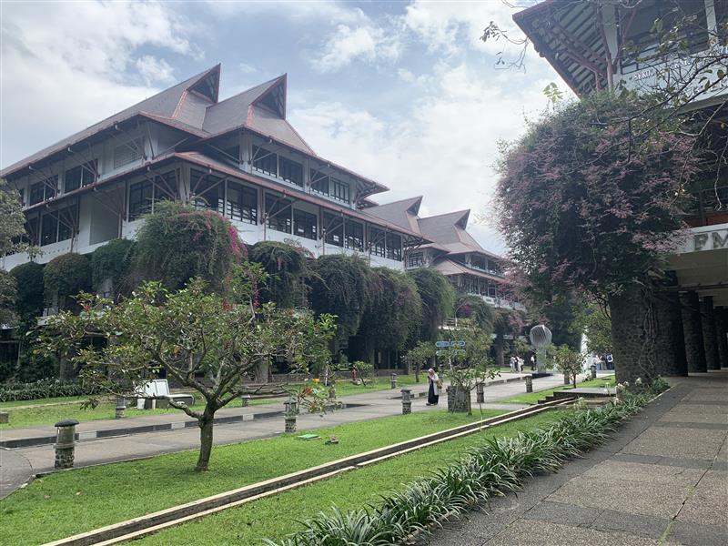 Traditional Indonesian-style campus buildings with steep multi-tiered roofs and vine-draped balconies overlooking a landscaped walkway and green lawn where a few students are walking.