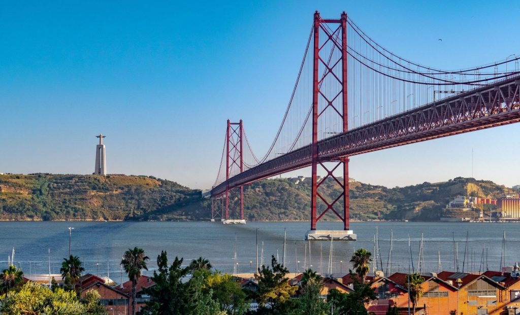 A red suspension bridge spans over a calm river with a large statue on a hill in the background. The foreground shows a marina with boats and palm trees.