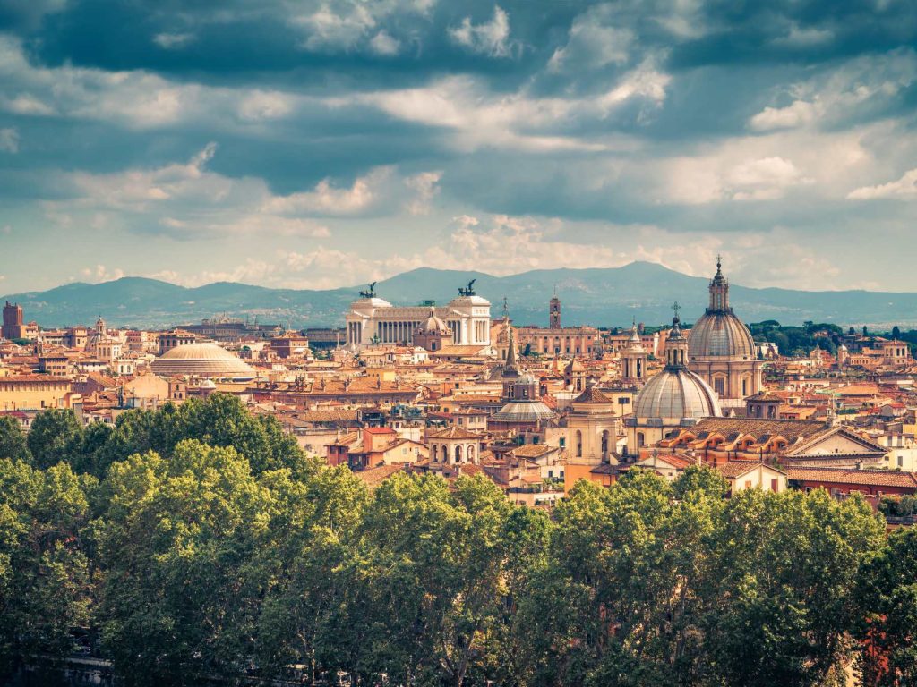 Scenic aerial view of Rome with domes, historic rooftops, and the Altare della Patria, set against tree-lined foreground and distant mountains.