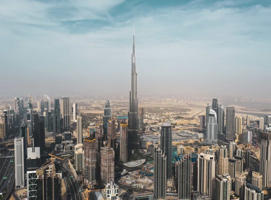 Daytime aerial view of downtown Dubai featuring the Burj Khalifa rising prominently above the dense cityscape of high-rise buildings and construction sites, under a partly cloudy sky. The image highlights the urban density and vertical architecture of the city.