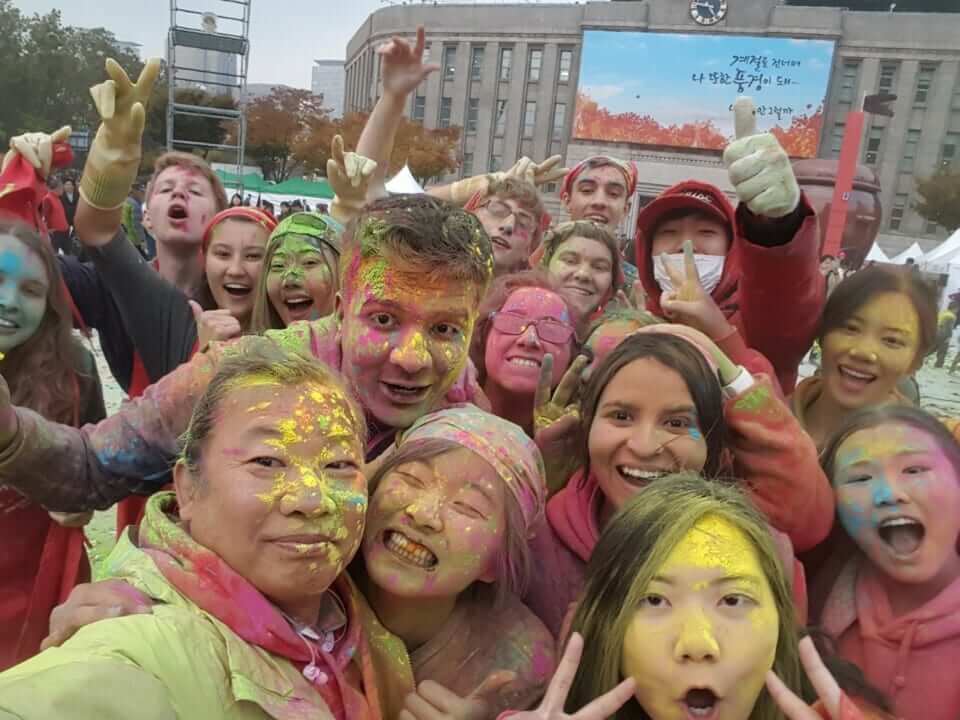 A diverse group of people, faces covered in vibrant powder, smile and pose for a joyful outdoor selfie during a colorful festival. The mood is lively and festive.