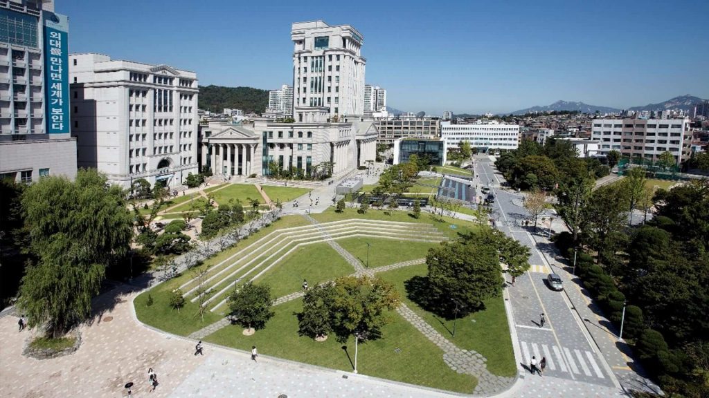 Aerial view of a university campus with modern and classical buildings surrounding a green park. Pathways and neatly trimmed trees create a serene atmosphere.