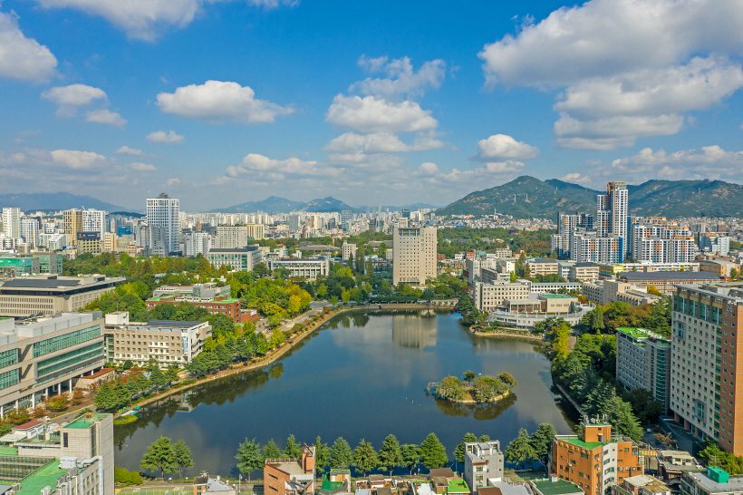 A breathtaking aerial view of Konkuk University in Seoul, featuring a large lake, modern buildings, and surrounding mountains, showcasing an ideal environment for students who want to study in Seoul.