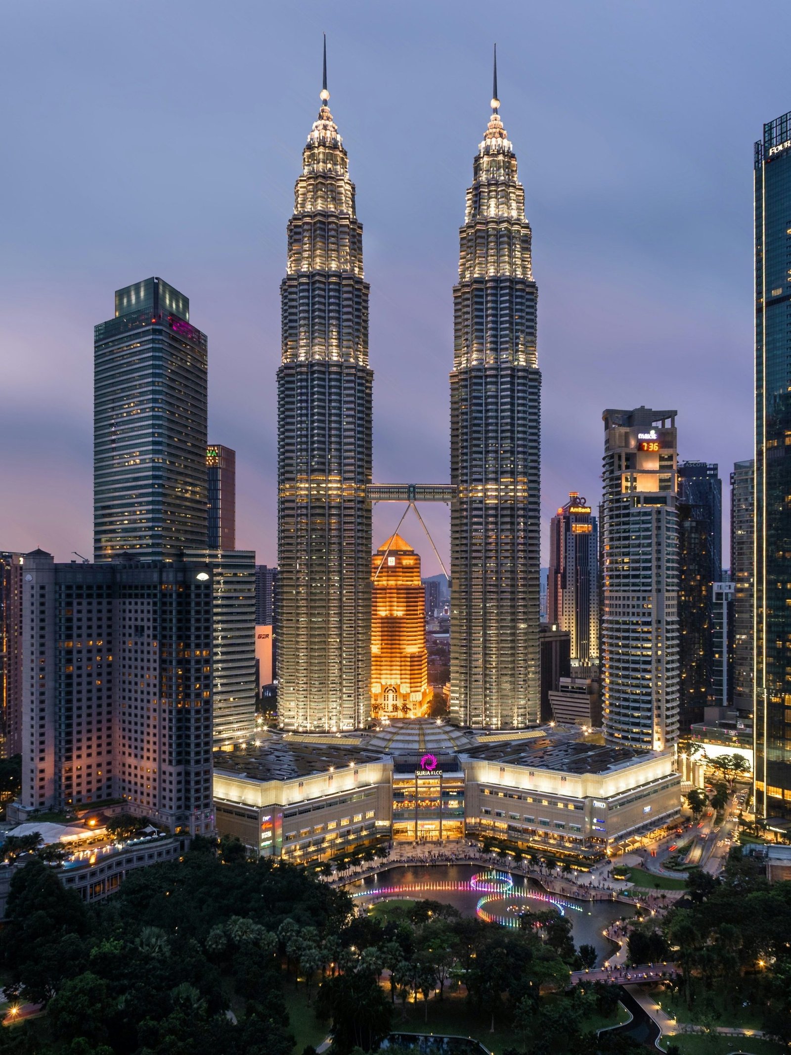 Evening cityscape of Kuala Lumpur featuring the illuminated Petronas Twin Towers surrounded by high-rise buildings. The foreground includes KLCC Park with trees and walkways, while soft purple twilight hues color the sky.