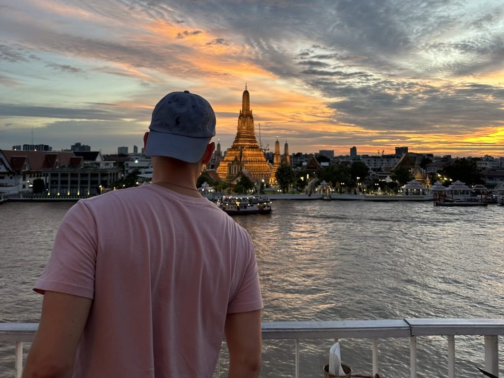 Noah, wearing a cap and pink t-shirt, stands at a railing looking across the Chao Phraya River at the illuminated Wat Arun temple in Bangkok, Thailand, under an orange sunset sky.