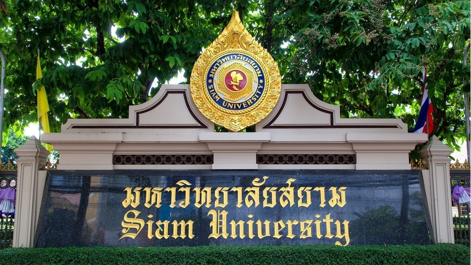 A sign for Siam University stands amid lush greenery. The logo is prominently displayed above, and text reads "Siam University" in gold on a dark stone background.